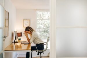 young woman seated at her desk in home office with her head in her hands struggling with emotional habits and addiction