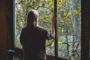 woman standing alone at window and staring out into the brightly lit woodlands wondering how to overcome social anxiety