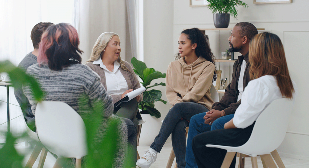 group of men and women seated in a circle discussing with a therapist what are the goals of group therapy